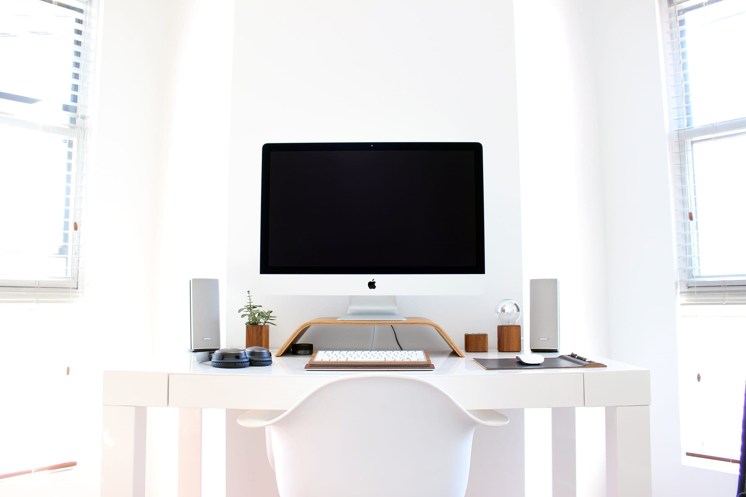Bureau blanc entre deux fenêtres, avec un écran surélevé sur un support en bois, un clavier centré et une chaise blanche
